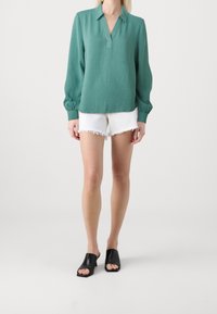 Blusa a maniche lunghe di colore teal con colletto, caratterizzata da una vestibilità comoda e polsini elasticizzati, abbinata a pantaloni corti bianchi sfilacciati e scarpe nere open-toe.