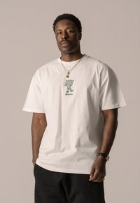 White cotton t-shirt with a loose fit, featuring a green illustration on the front. Model wears layered gold necklaces and dark pants.