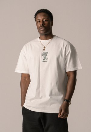 Witte katoenen t-shirt met een losse pasvorm, voorzien van een groene illustratie aan de voorkant. Model draagt gelaagde gouden kettingen en donkere broeken.