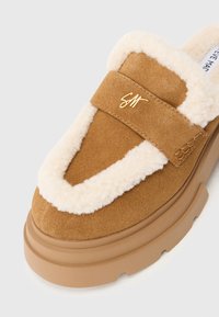 Scarpa slip-on in suede marrone con un morbido bordo in shearling crema, dotata di una fascia anteriore e di una robusta suola in gomma marrone. Dettaglio del logo oro presente.