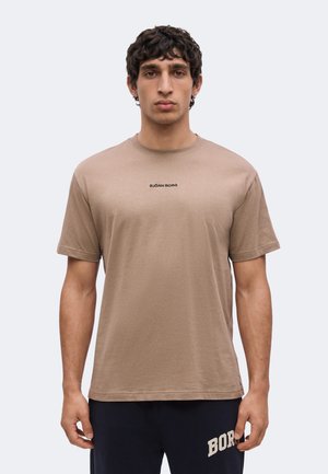 Jonge man met kort donker krullend haar, die een beige Björn Borg t-shirt en een donkere broek draagt, staand tegen een eenvoudige lichte achtergrond.