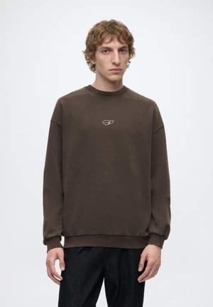 Ung mand med krøllet hår iført en brun sweatshirt med et lille logo på brystet og mørke bukser, står mod en ensfarvet baggrund.