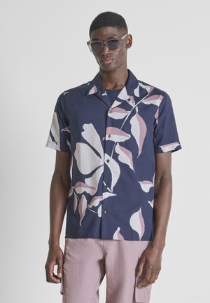 Jonge man draagt een donkerblauw overhemd met korte mouwen met een groot wit en roze bloemenpatroon, lichtroze cargobroek en een zonnebril.