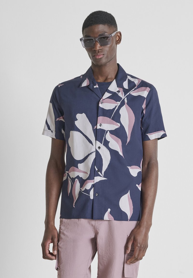 Joven con camisa azul oscuro de manga corta con botones y un gran estampado floral blanco y rosa, pantalones cargo rosa claro y gafas de sol.