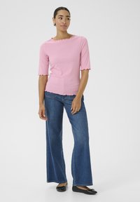 Roze geribbelde top met een golvende halslijn en mouwen, gecombineerd met een wijde blauwe jeans. Model draagt zwarte platte schoenen en staat voor een neutrale achtergrond.
