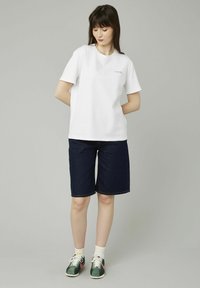 T-shirt blanc à manches courtes, bermuda en denim foncé, baskets vertes avec des accents roses, et chaussettes à rayures jaunes. Design simpliste et décontracté.