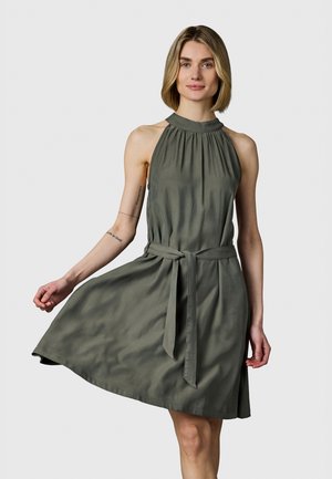 Kvinne iført ermeløs olivengrønn halterneck-kjole med midjebånd, holder skjørtet lett utstrakt, foran ensfarget lys grå bakgrunn.