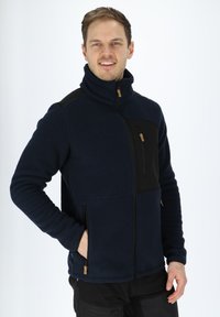 Giacca in fleece blu navy con accenti neri, colletto alto, tasca a zip sul petto e tasche laterali, con tessuto testurizzato e zip di colore beige.