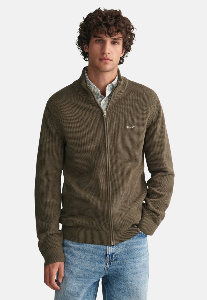 Maglione zip-up in verde oliva, tessuto texturizzato con polsini e orlo a costine. Presenta un piccolo logo sull'area sinistra del petto. Indossato sopra una camicia con collare.