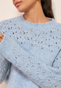 Svetlomodrý pletený sveter s textúrovaným, otvoreným čipkovým vzorom. Rebrované manžety a výstrih dodávajú detail mäkkej, nadýchanej tkanine.