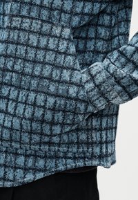 Μακρυμάνικο πουκάμισο με μπλε και μαύρο καρό, με μπροστινή τσέπη και μανσέτα με κουμπιά, φορεμένο με μαύρο παντελόνι.