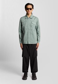 Lichtgroene button-up jas met twee borstzakken, gecombineerd met losse zwarte cargobroeken en zwarte sneakers. Gladde stof, relaxte fit.