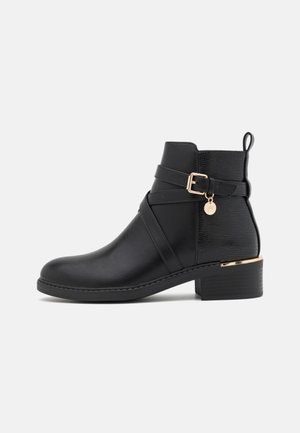 Black ankle boot made of smooth leather with a textured back panel. Features a gold-tone buckle and a small charm on the strap. Block heel.
