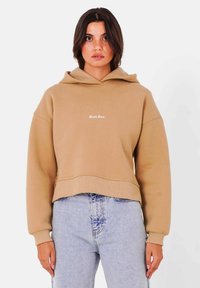 Sudadera beige recortada hecha de una tela suave, con un bolsillo canguro y capucha con cordón. Presenta un logo de texto blanco en el pecho.