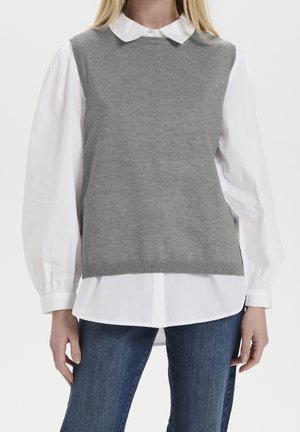 Femme portant un gilet sans manches en tricot gris sur un chemisier blanc à manches longues bouffantes et un jean bleu, se tenant devant un fond uni.