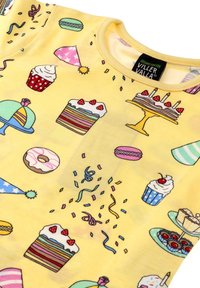 Keltainen kangas, jossa on erilaisia herkkuja: muffinssit, kakut, donitsit ja juhlahatut, joissa on värikkäitä kuvioita ja leikkisiä yksityiskohtia.