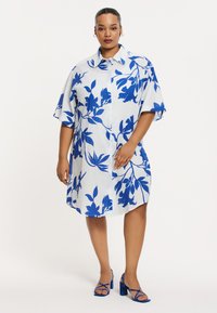 Blau-weißes, floral bedrucktes Oversized-Hemdkleid mit kurzen Ärmeln, Knopfleiste und lässigem Schnitt. Kombiniert mit blauen Riemchensandalen mit Blockabsatz.