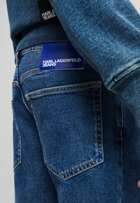 Jeansi din denim de culoare albastru mediu, cu o textură netedă. Au un buzunar din spate cusut și o etichetă albastră cu inscripția "KARL LAGERFELD JEANS."