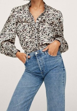 Woman wearing light blue high-waist jeans and a long-sleeve floral blouse with ruffle details, hands in jean pockets.