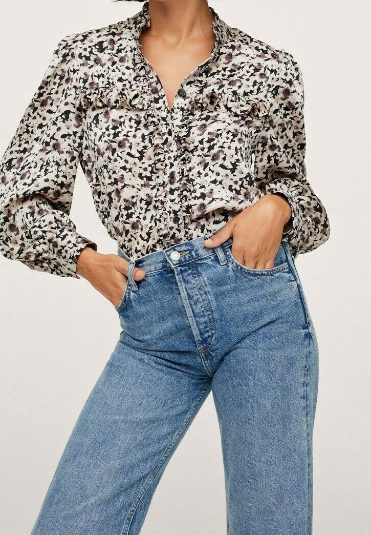 Femme portant un jean taille haute bleu clair et un chemisier à manches longues à imprimé floral avec des détails à volants, mains dans les poches du jean.