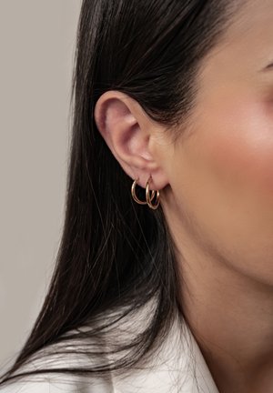 Nahaufnahme eines Frauenohres mit zwei kleinen goldenen Creolen und langen dunklen Haaren vor neutralem Hintergrund.