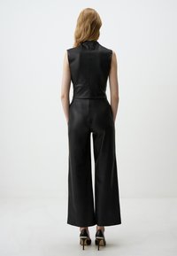 Zwart mouwloos jumpsuit van glad leer met een hoge halslijn, strakke lijfje en wijde broekspijpen, gecombineerd met zwarte hoge hakken.