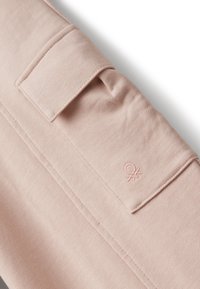 Světle růžový látkový rukáv s přišitým obdélníkovým klopovým kapsou a malým vyšitým logem značky blízko manžety.