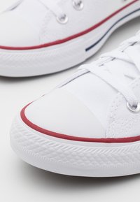 Gros plan de baskets en toile blanches avec des lacets blancs, une bordure rouge autour de la semelle en caoutchouc, et des œillets métalliques.