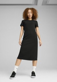 Vestito nero, fino al ginocchio, aderente con maniche corte. Texture liscia, design minimale. Abbinato a sneakers nere e calze bianche.