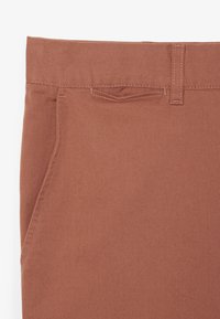 Pantaloni di cotone marroni con cintura, tasche frontali e texture diagonale. Presentano cuciture a contrasto lungo i bordi.