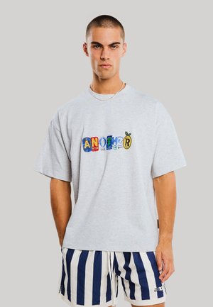 Ung mand med kort hår iført en lysegrå oversized t-shirt med farverig tekst og marineblå og hvide lodrette stribede shorts.