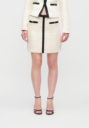 Kobieta ubrana w kremową teksturowaną spódnicę i dopasowaną marynarkę z czarną lamówką, zestawione z czarnymi sandałami na wysokim obcasie z paskami.