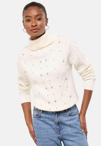 Krém színű, gyöngyház mintás kötött garbó, melyen rombusz minták, apró díszítések és bordázott mandzsetták találhatók, kék farmerrel párosítva.