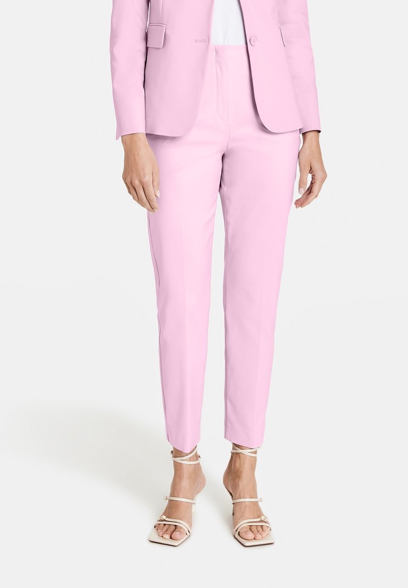 Femme portant un pantalon rose clair taillé sur mesure et un blazer assorti avec des sandales blanches à talons hauts à brides, debout sur un fond blanc.