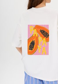 Witte t-shirt met korte mouwen, voorzien van een afbeelding van twee gehalveerde papaja's met zwarte zaden, en gele letters die "PAPA" spellen, tegen een achtergrond van roze strepen.