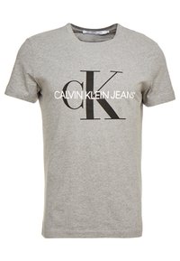 Grå bomullströja med rund halsringning som har en stor svart "CK"-logotyp och texten "CALVIN KLEIN JEANS" i vitt. Kort ärm, avslappnad passform.