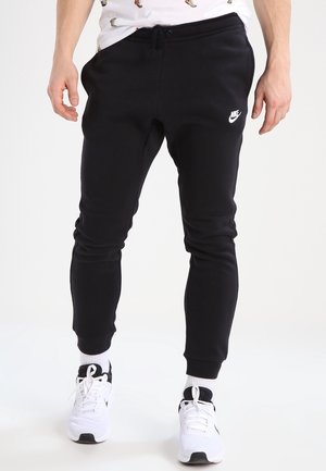 Man draagt zwarte Nike joggingbroek, witte sportsokken en witte sneakers, staand tegen een effen witte achtergrond.