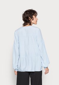 Blusa azul clara de manga longa com um corte descontraído, apresentando um design em camadas e punhos franzidos. Textura suave, decote redondo e estilo minimalista.