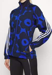 Chaqueta de cremallera azul con estampado floral y cuello alto, que presenta rayas blancas en las mangas y un acento dorado con el logo. Fabricada con material suave.