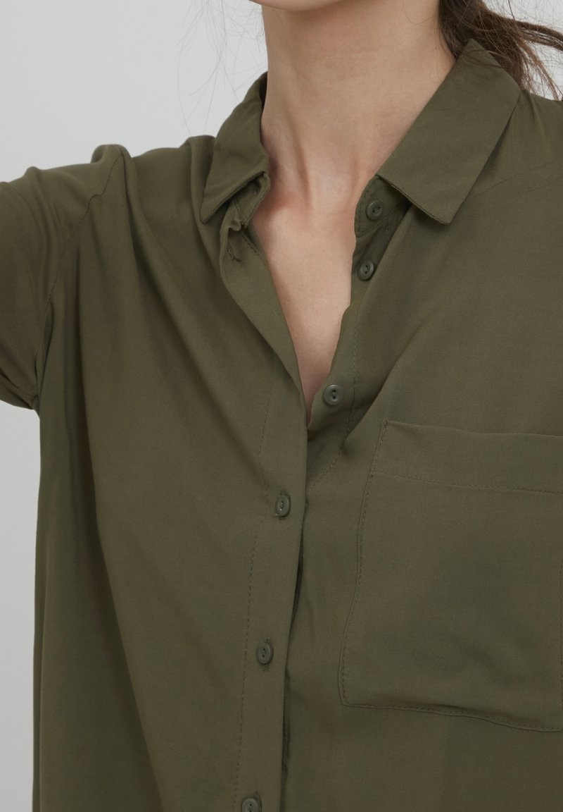 Olivgrön skjorta med knappar som har krage, kort ärm och bröstficka. Slät textur med tygknappar.