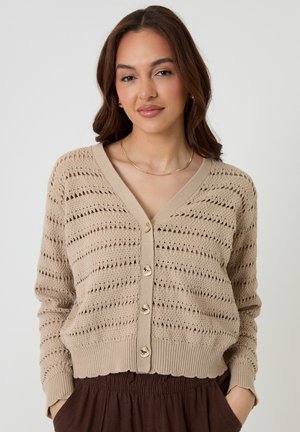 Vrouw met bruin haar, gekleed in een beige gebreid vest met knopen en donkerbruine broek, staande met handen in de zakken tegen een effen achtergrond.