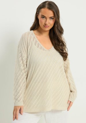 Kvinde med langt brunt hår iført en løs cremefarvet strikket sweater med diagonale åbne strikkemønstre og hvide bukser foran en ensfarvet baggrund.