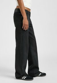 Jeans de pierna ancha negros con una textura de denim suave, que cuentan con cinco bolsillos y una cinturilla negra con logotipo en relieve. Combinados con zapatillas negras.