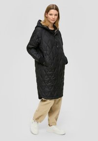 Schwarze gepolsterte Jacke mit einem Rautenmuster, ausgestattet mit einer Kapuze, Reißverschluss vorne und seitlichen Taschen. Getragen über beigen Hosen und weißen Sneakern.