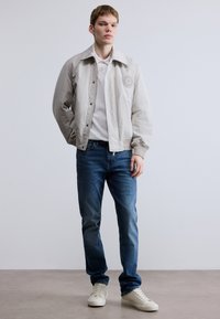 Svijetlosiva bomber jakna s teksturiranim finišem i logotipom sprijeda, bijela polo majica, plave traperice i kremaste tenisice, stojeći na neutralnoj pozadini.