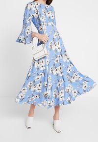 Floral maxi dress in light blue fabric with white and grey flowers, featuring bell sleeves. Paired with white mules and a small white crossbody bag.