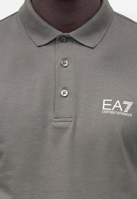 Gros plan d'un polo gris avec trois boutons et le logo blanc EA7 Emporio Armani sur la partie droite de la poitrine.
