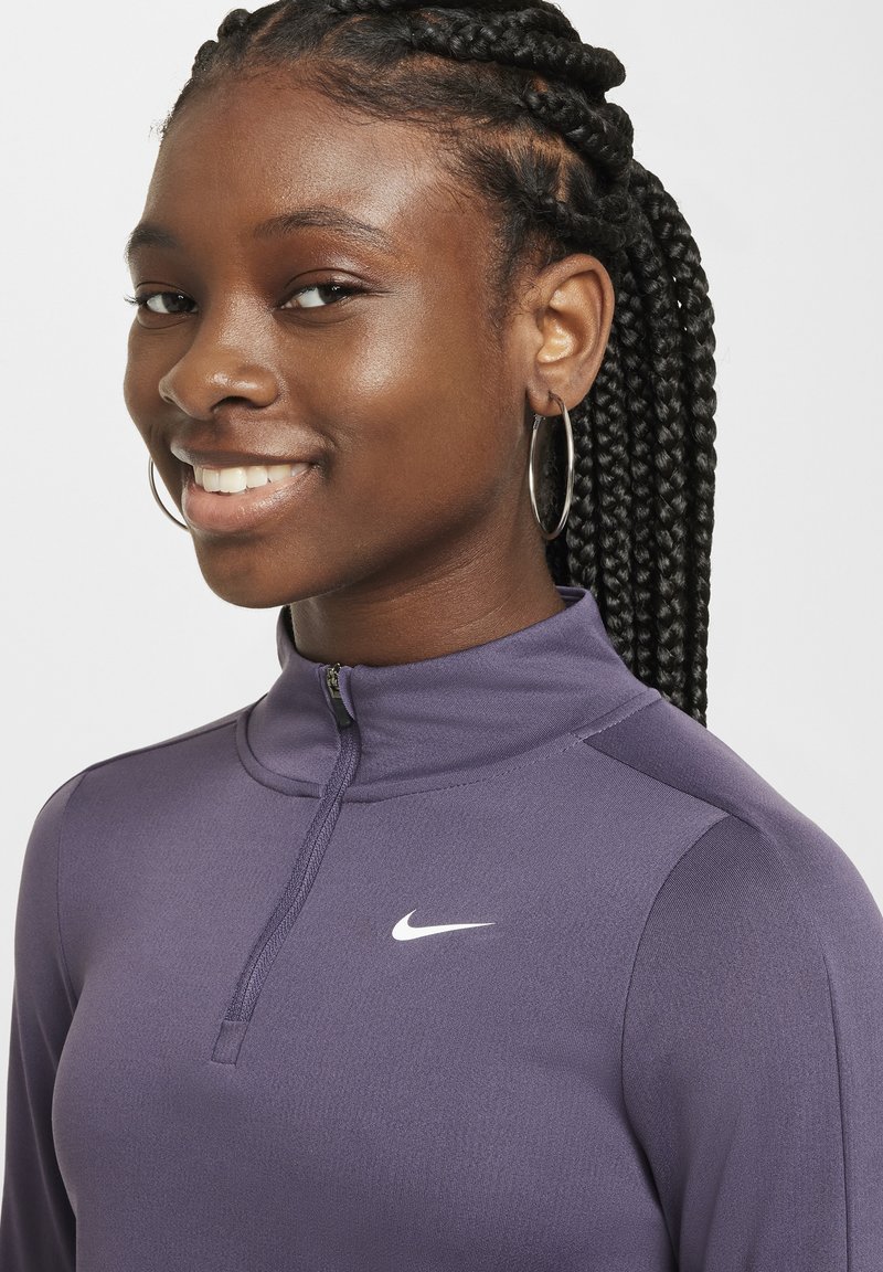 Maglietta sportiva a maniche lunghe di colore viola con zip a quarti e logo Nike sul petto. Tessuto liscio, design aderente. Capelli intrecciati e orecchini a cerchio.