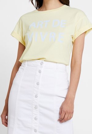 Femme portant un t-shirt jaune pâle avec le texte "ART DE VIVRE" et une jupe blanche taille haute à boutons.