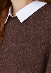 Brun textured tröja med en vit krage. Nära vy som framhäver stickmönstret och den kontrasterande kragen.